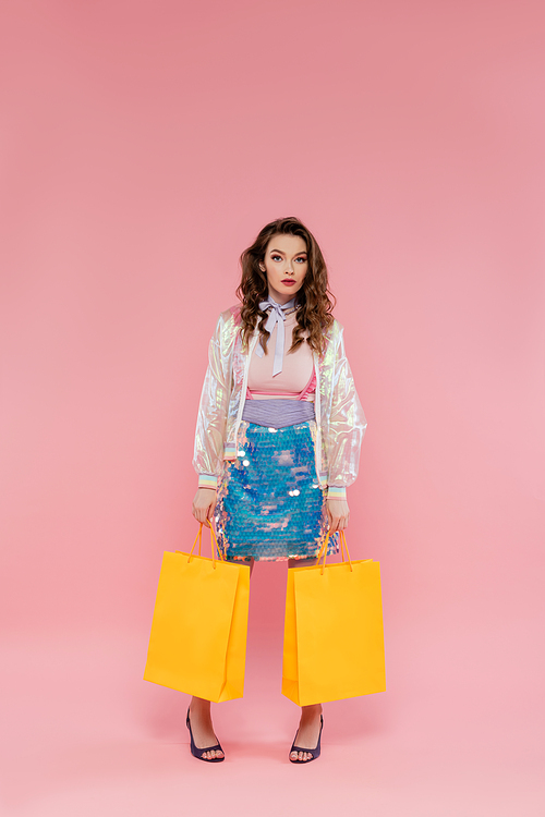 attractive woman carrying shopping bags on pink background, concept photography, consumerism, stylish outfit, young model in skirt with sequins and transparent jacket looking at camera