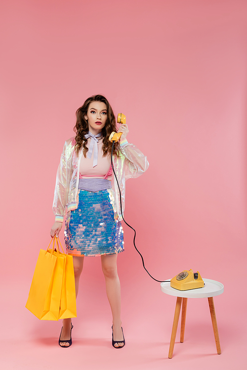 beautiful young woman carrying shopping bags, talking on retro telephone, standing like a doll on pink background, concept photography, doll pose, model in skirt with sequins and transparent jacket