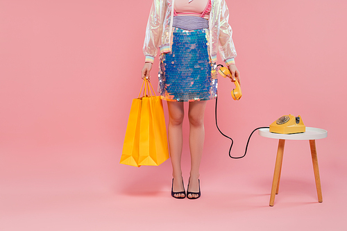 conceptual photography, cropped view of young woman carrying shopping bags and holding retro telephone, standing on pink background, phone call, vintage telephone, housewife concept