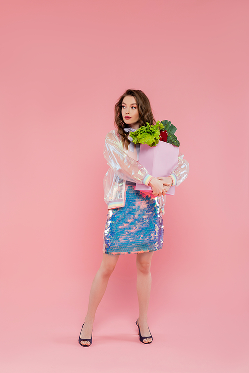 housewife concept, attractive young woman carrying grocery bag with vegetables, model with wavy hair standing on pink background, conceptual photography, home duties, stylish wife