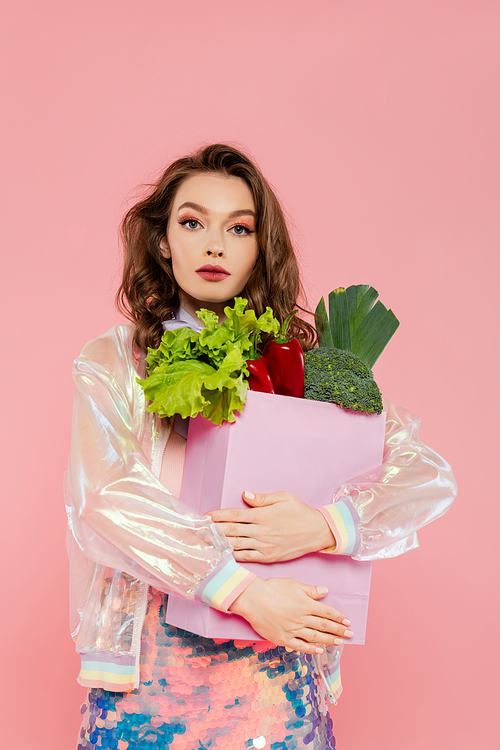 housewife concept, beautiful young woman carrying grocery bag with vegetables, model with wavy hair standing on pink background, conceptual photography, home duties, stylish wife, portrait