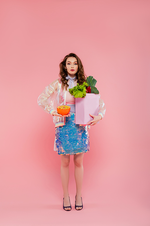 housewife concept, doll like, attractive young woman carrying grocery bag with vegetables, model with wavy hair on pink background, conceptual photography, home duties, stylish wife