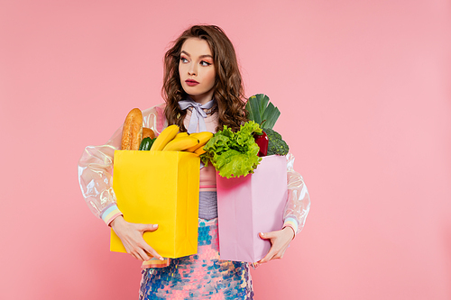 housewife concept, attractive young woman carrying grocery bags with vegetables and bananas, model with wavy hair on pink background, conceptual photography, home duties, stylish wife