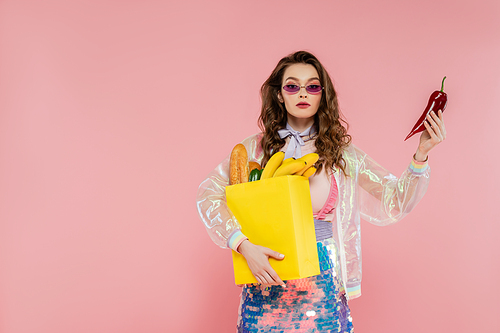 housewife concept, attractive young woman in sunglasses carrying paper bag with groceries and holding red pepper, posing like a doll on pink background, conceptual photography, home duties