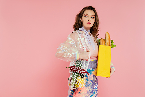 housewife concept, beautiful young woman holding reusable mesh bag with groceries, stylish wife doing daily house duties, standing on pink background, looking away, role play