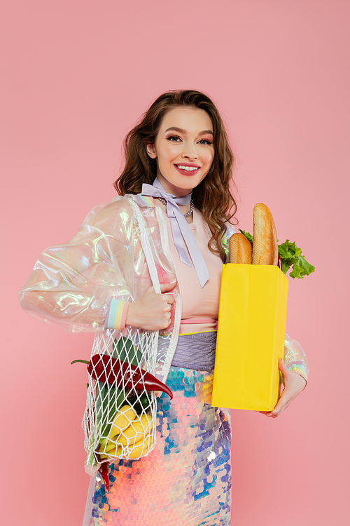 housewife concept, happy young woman holding reusable mesh bag with groceries, stylish wife doing daily house duties, standing on pink background, looking at camera, role play