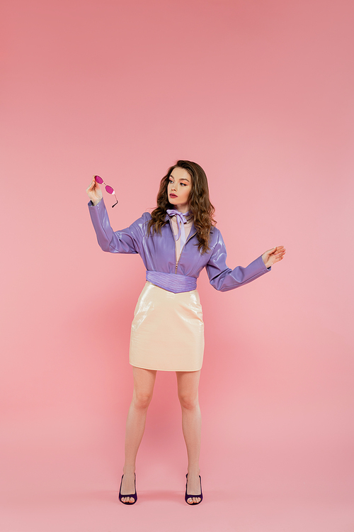 glamour, beautiful young woman gesturing and looking at sunglasses, acting like a doll, fashionable outfit, model in purple jacket and skirt, pink background, studio shot, conceptual, full length