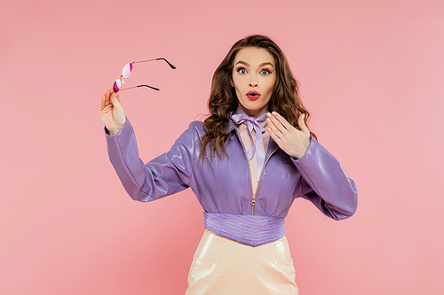 glamour, surprised young woman gesturing and looking at camera, holding sunglasses, fashionable outfit, model in purple jacket and skirt standing on pink background, studio shot, acting like a doll