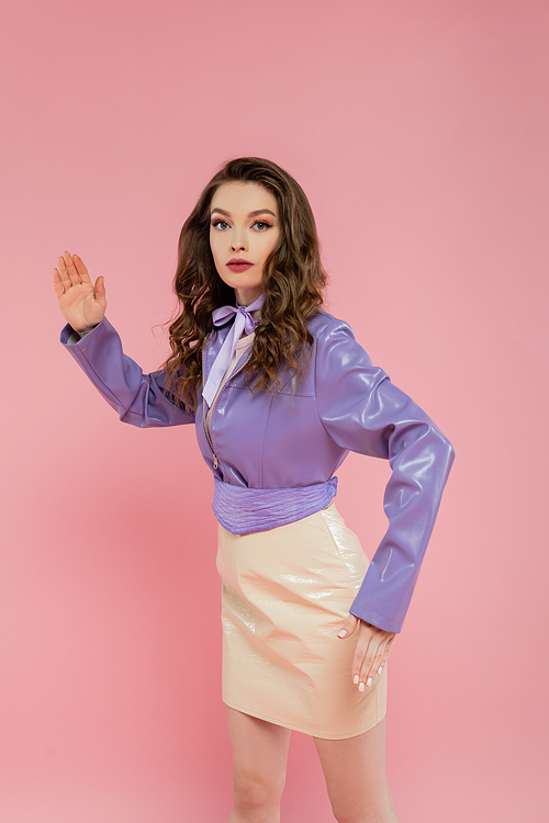 doll concept, beautiful young woman with wavy hair gesturing and looking at camera, trendy outfit, brunette model in purple jacket posing on pink background, studio shot, conceptual