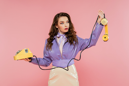 doll pose, beautiful young woman with wavy hair looking at handset while holding yellow retro phone, brunette model in purple jacket posing on pink background, studio shot