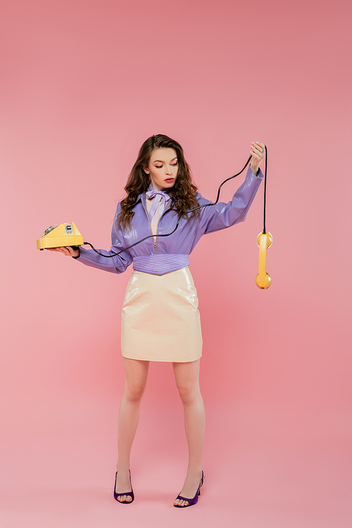 doll pose, beautiful young woman with wavy hair looking at handset while holding yellow retro phone, brunette model in purple leather jacket posing on pink background, studio shot, full length