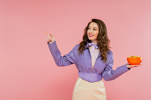 concept photography, cheerful woman with wavy hair pretending to be a doll, pointing away, holding bowl with corn flakes, eating tasty breakfast, posing on pink background, stylish purple jacket