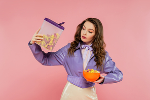 concept photography, brunette woman with wavy hair pretending to be a doll, holding container with corn flakes, tasty breakfast, posing on pink background, stylish purple jacket