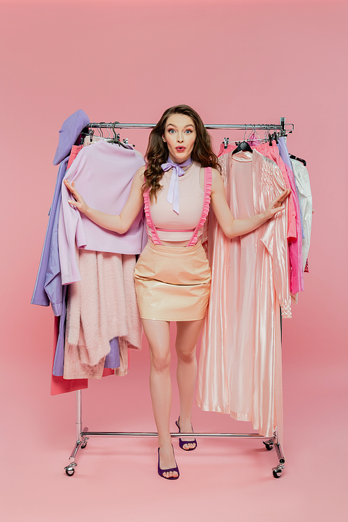 consumerism, amazed young woman with brunette wavy hair standing near rack with clothes, wardrobe selection concept, personal style, fashion and trends, beautiful model, emotional
