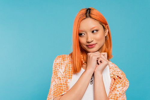 adorable, cheerful young asian woman with dyed red hair smiling and holding hands near face on vibrant blue background, pleased, generation z, casual attire, looking away, young culture