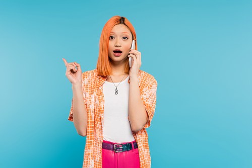 phone call, advertisement, young asian woman with dyed hair pointing with finger and talking on smartphone on blue background, mobile phone, youth culture, digital age, generation z, surprised face