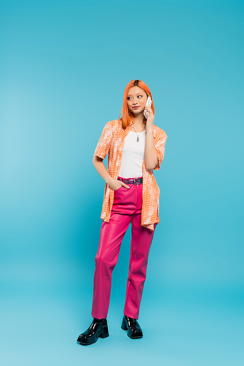 hand in pocket, smiling and young asian woman with dyed hair standing in casual attire and talking on smartphone on blue background, digital age, generation z, modern style, full length