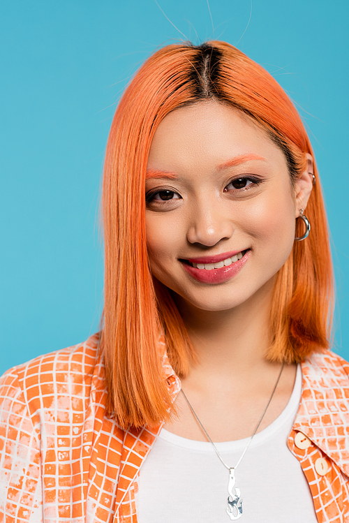 portrait, young asian woman with short and dyed hair, natural makeup and hoop earrings looking at camera on blue background, orange shirt, generation z, fashion, happy face, radiant smile
