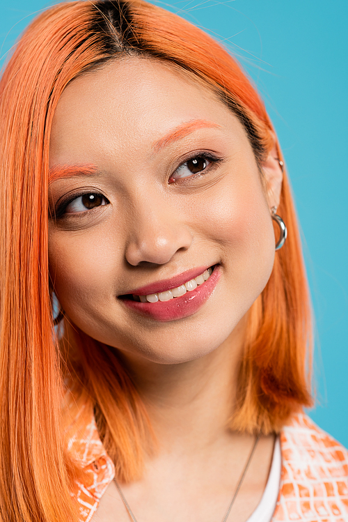 portrait, joyful asian woman with short and dyed hair, natural makeup and hoop earrings looking away on blue background, orange shirt, generation z, fashion, happy face, radiant smile