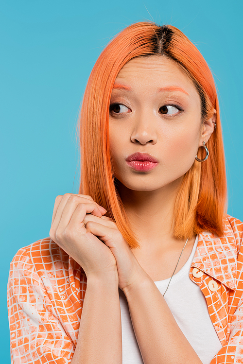 portrait, curious young asian woman with short and dyed hair, natural makeup and hoop earrings looking away on blue background, orange shirt, generation z, pouting lips, emotion