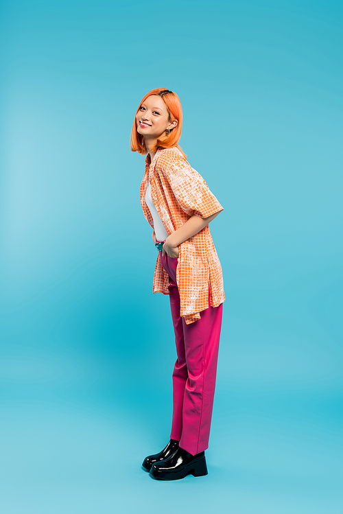 posing with hand in pocket, joyful and young asian woman with dyed hair standing in casual wear on blue background, looking at camera, pink pants, generation z, modern style, full length