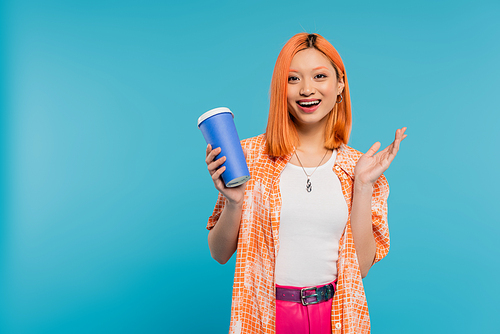 positivity, drink in hand, happy asian and young woman with red hair holding paper cup and looking at camera on blue background, casual attire, generation z, coffee culture, hot beverage, amazed