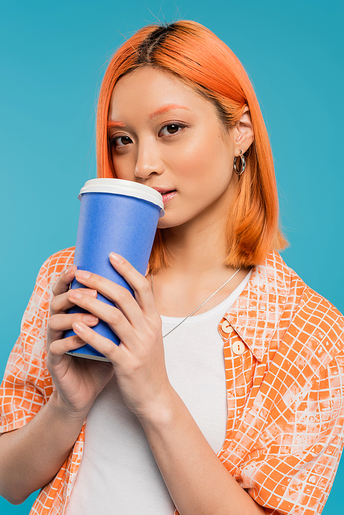 female model drinking coffee to go, asian and young woman with red hair holding paper cup and looking at camera on blue background, casual attire, generation z, coffee culture, hot beverage
