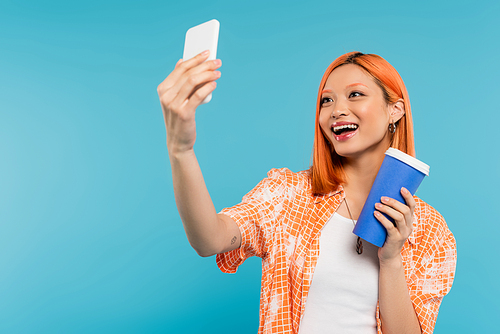 taking selfie, happy asian and young woman with red hair holding paper cup and using smartphone on blue background, casual attire, generation z, coffee culture, social media influencers