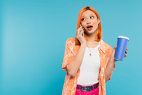 phone call, emotional asian and young woman with red hair holding takeaway drink and talking on smartphone on blue background, casual attire, generation z, coffee culture, amazed