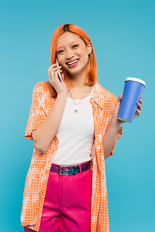 phone call, cheerful asian woman with red hair holding coffee to go in paper cup and talking on smartphone on blue background, casual attire, generation z, coffee culture, hot beverage