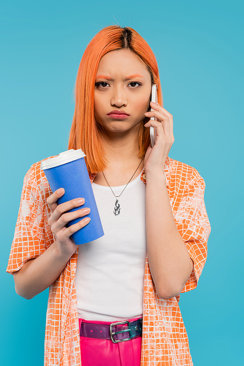 phone call, upset asian woman with red hair holding coffee to go in paper cup and talking on smartphone on blue background, casual attire, generation z, coffee culture, displeased, pouting lips