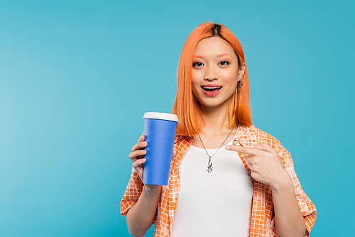 positivity, happy asian and young woman with red hair pointing at paper cup and looking at camera on blue background, casual attire, generation z, coffee culture, hot beverage, amazed