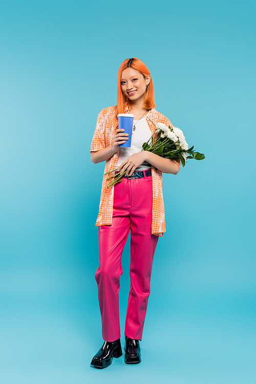 full length of cheerful asian and young woman with red hair holding coffee to go and bouquet of flowers on blue background, casual attire, generation z, coffee culture, hot beverage, paper cup