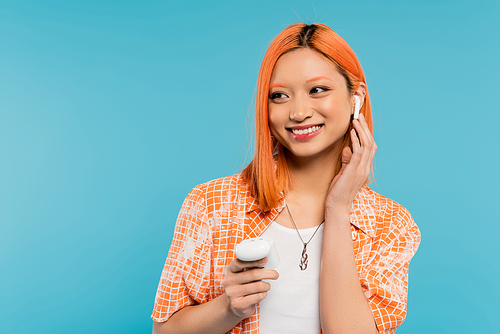 summer happiness, music lover, young and trendy asian woman with dyed hair, in orange shirt holding case, adjusting wireless earphone and looking away on blue background, generation z