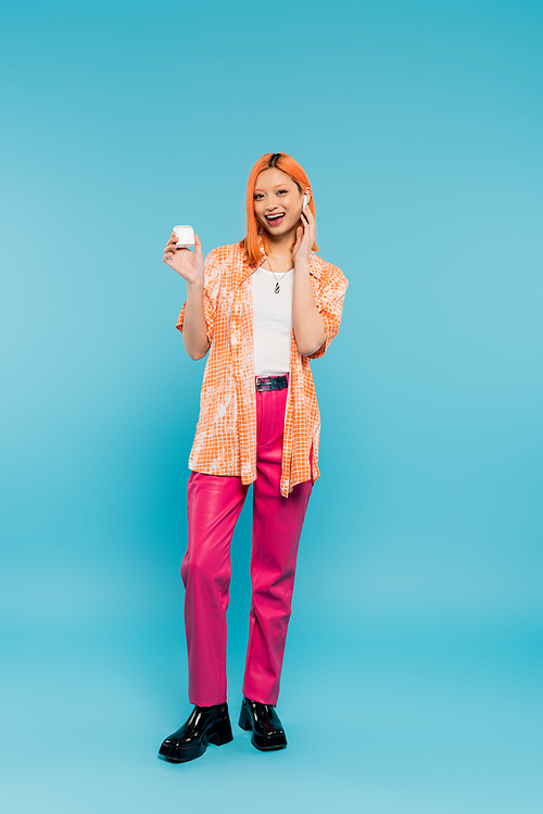 full length of happy and stylish asian woman with colored red hair, in orange shirt and pink pants holding earphone case and listening music while standing on blue background in studio