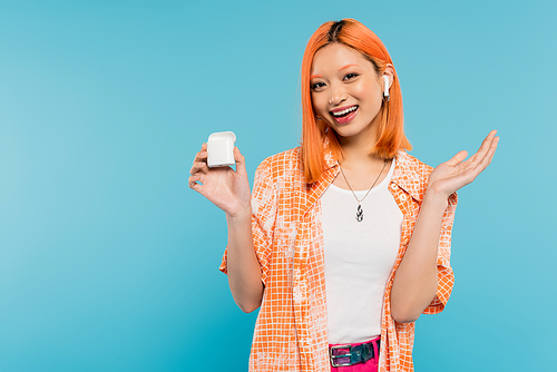 amazed asian woman with happy face and colored red hair wearing stylish orange shirt and listening music in wireless earphone while standing with case on blue background, youth culture