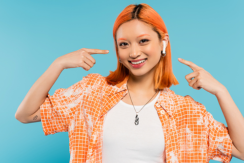 carefree summer, trendy asian woman with red hair and in orange blouse pointing with fingers at earphone, listening music and looking at camera on blue background, tattoo, generation z lifestyle