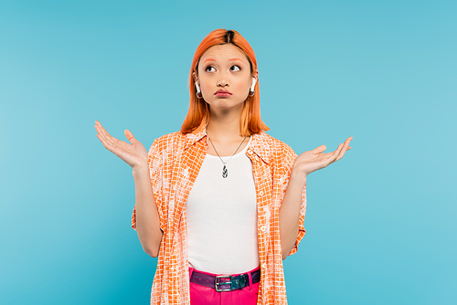 young and discouraged asian woman in orange shirt, with dyed red hair listening music in wireless earphone, showing shrug gesture and looking up on blue background, generation z