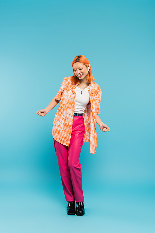 summer leisure, music lover, overjoyed asian woman in colorful clothes and wireless headphones on blue background, dyed hair, orange shirt, pink pants, gen z fashion