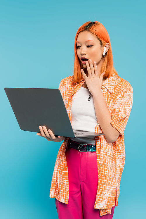 amazed asian woman with red colored red hair holding hand near open mouth and looking at laptop on blue background, youthful fashion, orange shirt, freelance lifestyle