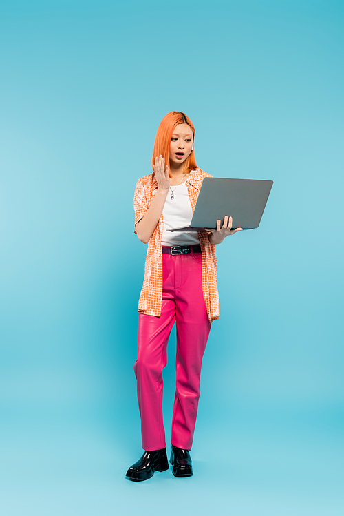 surprised emotion, video call, full length of redhead asian woman with open mouth, in orange shirt and pink pants waving hand near laptop on blue background, freelance lifestyle, generation z