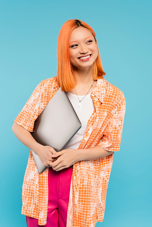 summer happiness, appealing asian woman with red hair and radiant smile holding laptop and looking away while standing in orange shirt on blue background, freelance lifestyle, generation z