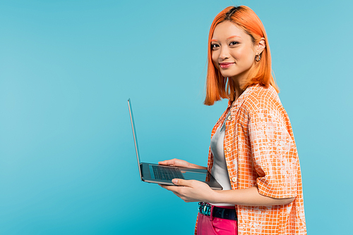 happy face, optimistic emotion, youthful asian woman with colored red hair, in orange shirt holding laptop and looking at camera on blue background, freelance lifestyle, generation z