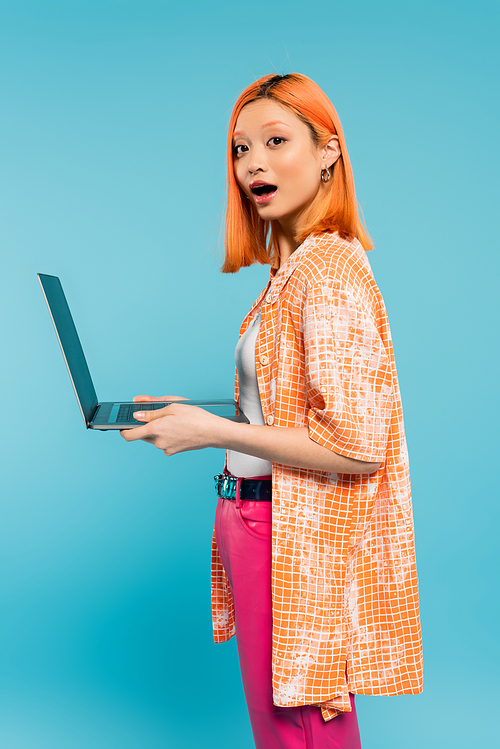 amazement, surprise, young asian woman with red colored red hair and open mouth holding laptop and looking at camera on blue background, youthful fashion, orange shirt, freelance lifestyle