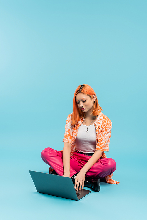 trendy asian woman in pink pants and orange shirt working on laptop while sitting with crossed legs on blue background, smiling, positive emotion, freelance lifestyle, generation z