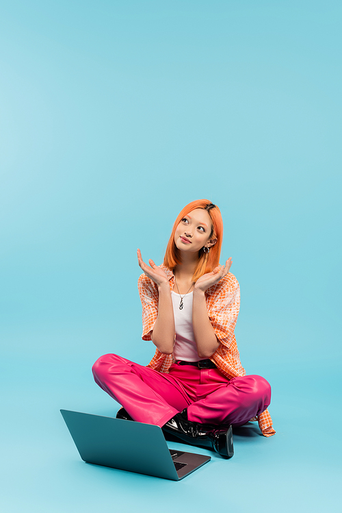 positive emotion, dreamy and smiling asian woman sitting with crossed legs near laptop on blue background, colored red hair, orange shirt, pink pants, freelance lifestyle, generation z