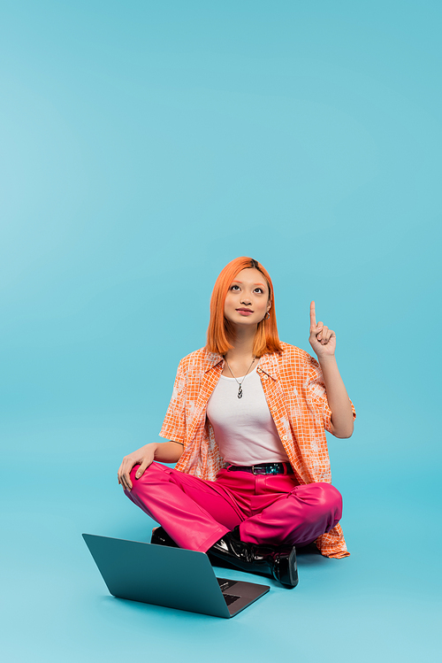 creative thinking, idea gesture, solution, redhead asian woman in orange shirt sitting with crossed legs near laptop, looking up and pointing with finger on blue background, freelance lifestyle