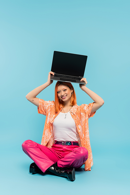 happiness, asian woman with cheerful face and red hair holding laptop with blank screen over head while sitting with crossed legs on blue background, tattoo, young freelancer, youth culture