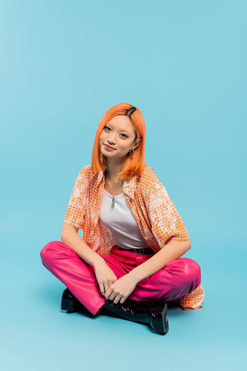 happy face, positive emotion, redhead and stylish asian woman sitting with crossed legs and looking at camera on blue background, orange shirt, pink pants, summer vibes