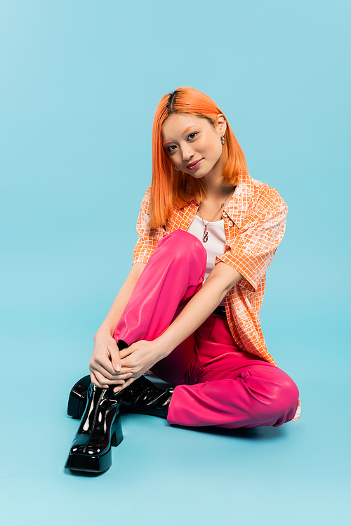 vibrant and happy personality, full length of young and trendy asian woman sitting and smiling at camera on blue background, colored red hair, pink pants, orange shirt, youth culture
